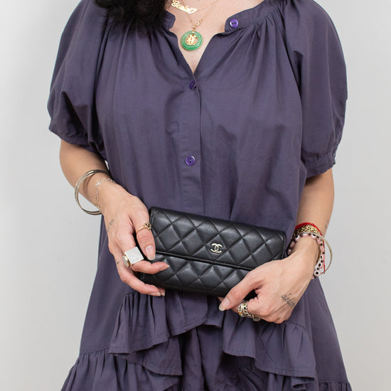 A woman wearing a short, dark purple puff-sleeve blouse with a ruffled hem, holding a black quilted clutch bag with a silver logo.