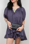 A woman wearing a short, dark purple puff-sleeve blouse with a ruffled hem, holding a black quilted clutch bag with a silver logo.