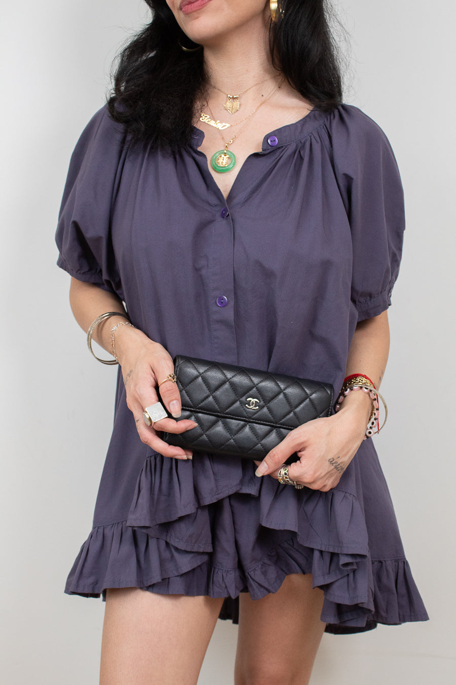 A woman wearing a short, dark purple puff-sleeve blouse with a ruffled hem, holding a black quilted clutch bag with a silver logo.
