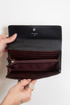 An open Chanel wallet in black leather with a burgundy interior, held by two hands against a plain white background.