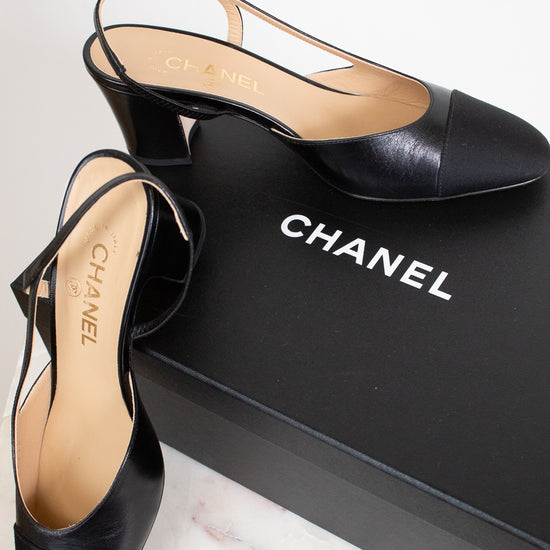 A pair of Chanel black leather slingback mule heels with a block heel and pointed toe, displayed next to their black Chanel shoebox on a light marble surface.