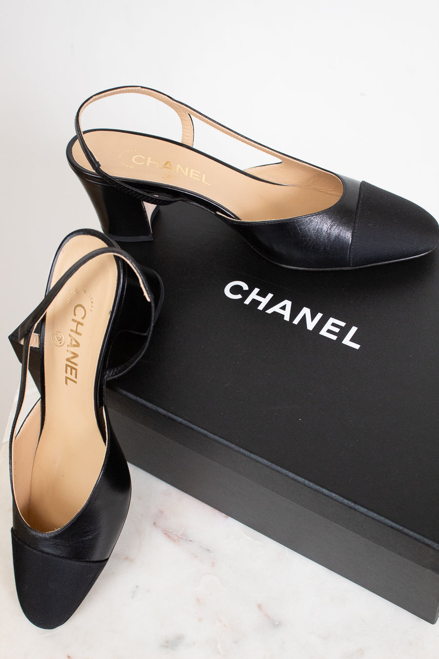 A pair of Chanel black leather slingback mule heels with a block heel and pointed toe, displayed next to their black Chanel shoebox on a light marble surface.