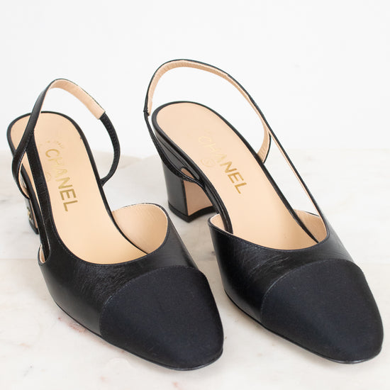 A pair of Chanel black leather slingback pumps with a low block heel and a contrasting black toe cap, displayed on a light marble surface against a white background.