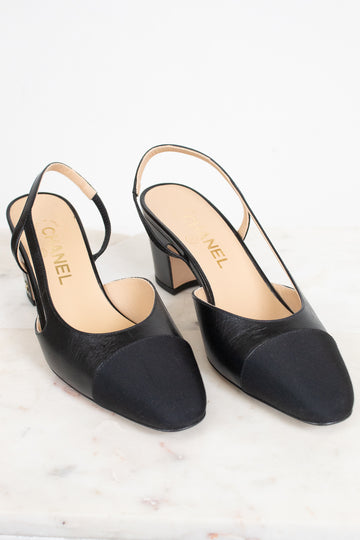 A pair of Chanel black leather slingback pumps with a low block heel and a contrasting black toe cap, displayed on a light marble surface against a white background.