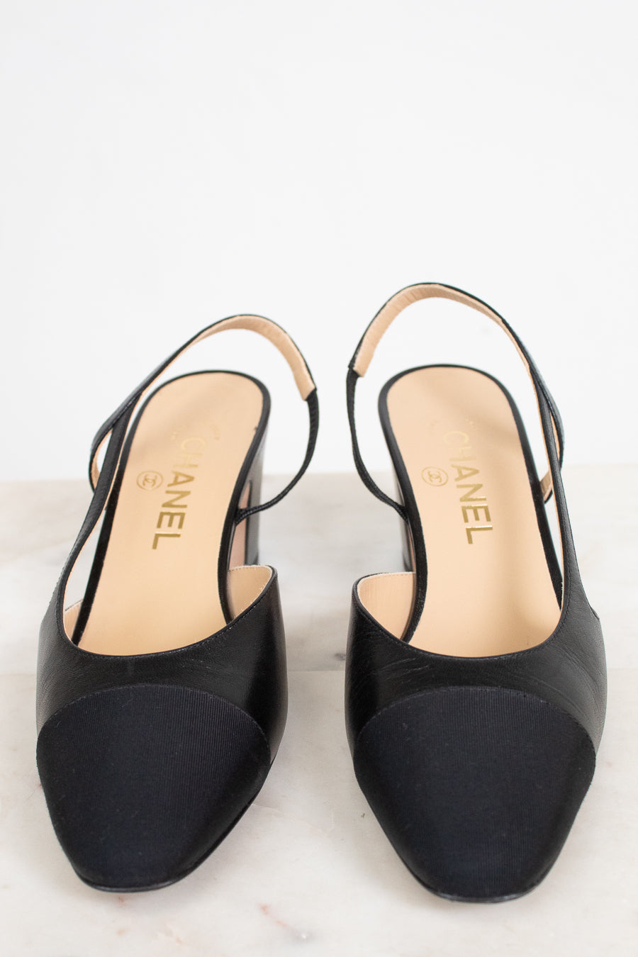 A pair of Chanel slingback high heels in black leather with a contrasting black suede toe cap, shown on a light marble surface against a white background.