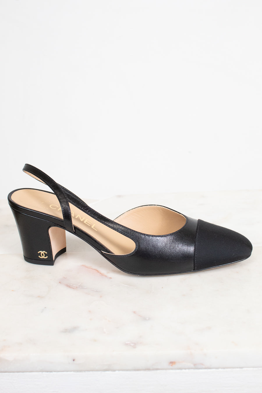 A single Chanel black leather slingback pump with a cap toe and block heel, resting on a white marble surface against a white background.