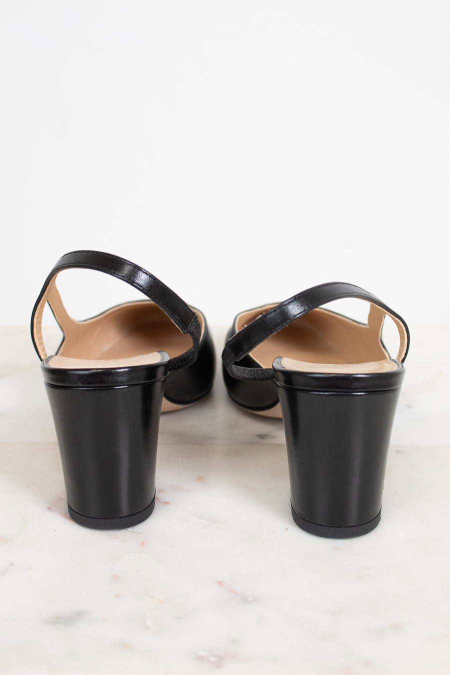 A pair of black leather slingback block-heel sandals shown from the back, resting on a light marble surface against a white background.