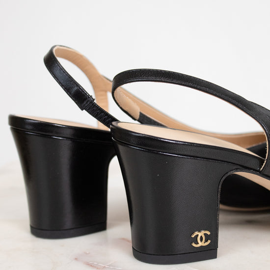 A pair of black leather Chanel slingback block-heel sandals with a gold interlocking CC logo on the heel, displayed on a light marble surface against a white background.