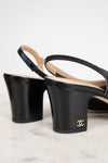 A pair of black leather Chanel slingback block-heel sandals with a gold interlocking CC logo on the heel, displayed on a light marble surface against a white background.