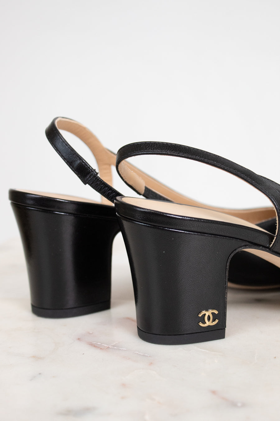 A pair of black leather Chanel slingback block-heel sandals with a gold interlocking CC logo on the heel, displayed on a light marble surface against a white background.