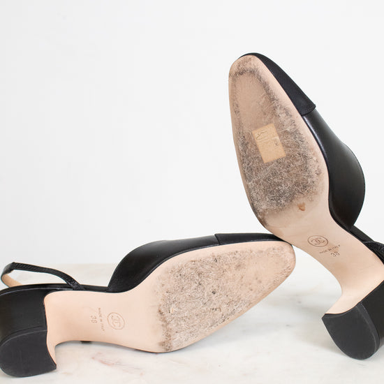 Black leather slingback high-heeled sandals shown from the sole, resting on a white marble surface against a white background.