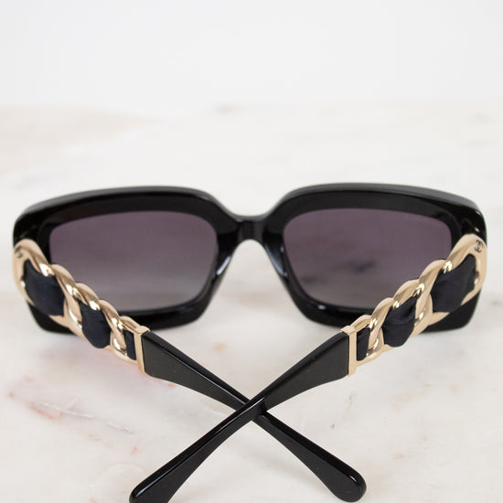 Black sunglasses with dark lenses and decorative gold-tone chain details on the arms, resting on a light marble surface against a white background.