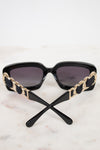 Black sunglasses with dark lenses and decorative gold-tone chain details on the arms, resting on a light marble surface against a white background.