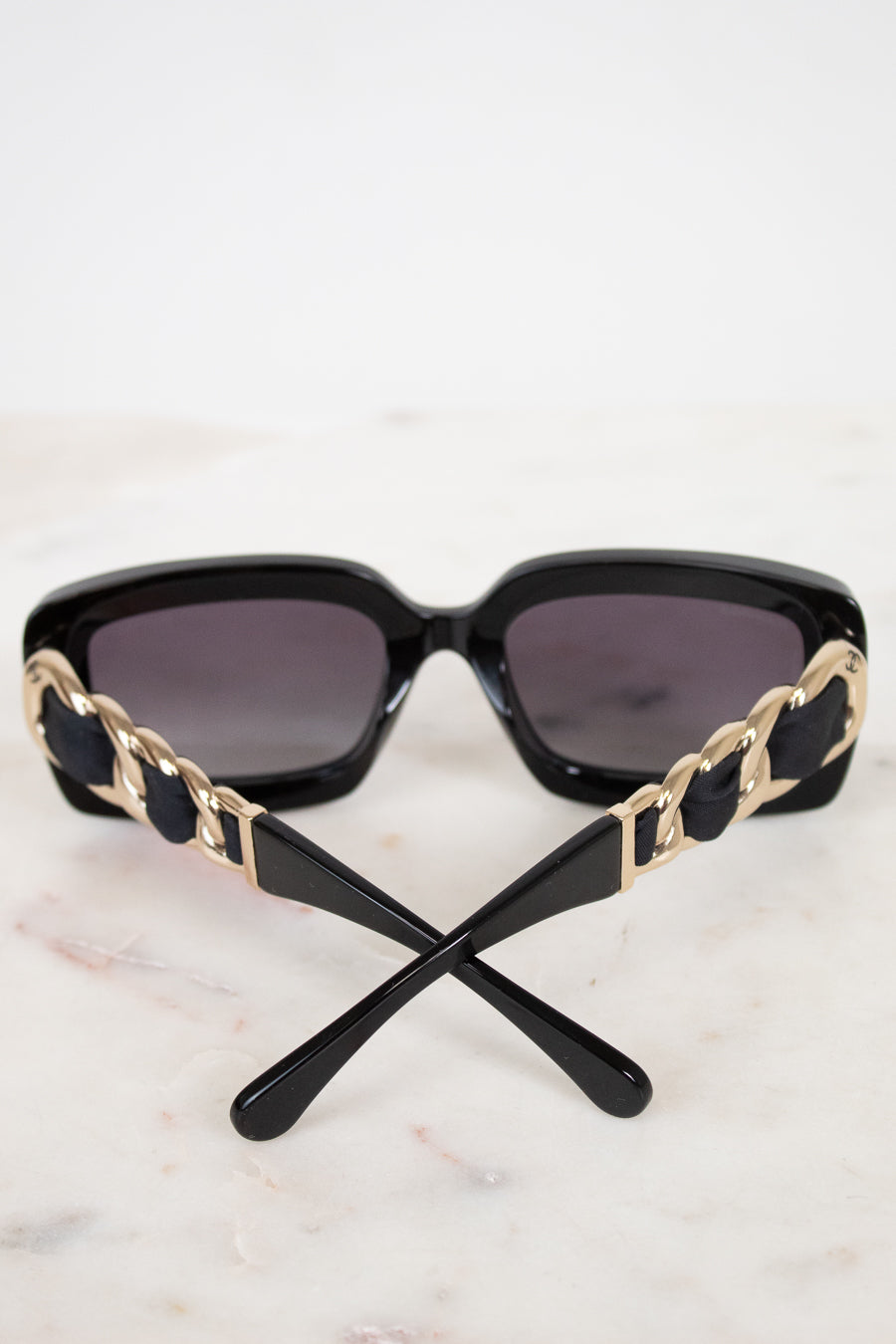 Black sunglasses with dark lenses and decorative gold-tone chain details on the arms, resting on a light marble surface against a white background.