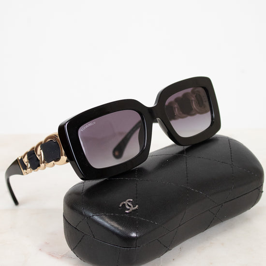 Chanel sunglasses with a black acetate frame, gold-tone chain detailing on the arms, and dark lenses, displayed on top of their signature black quilted leather case against a white marble background.