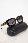 Chanel sunglasses with a black acetate frame, gold-tone chain detailing on the arms, and dark lenses, displayed on top of their signature black quilted leather case against a white marble background.