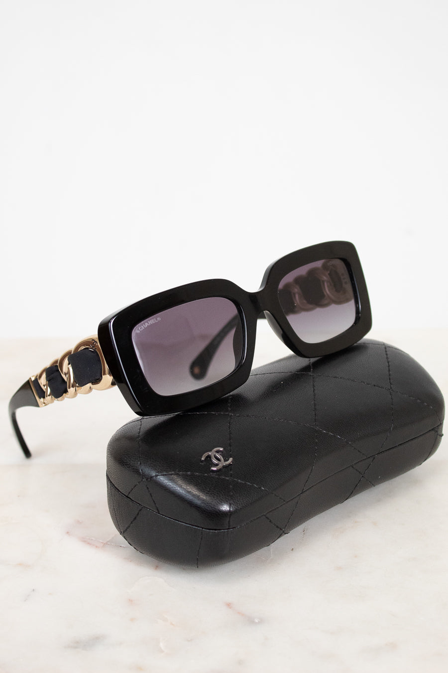 Chanel sunglasses with a black acetate frame, gold-tone chain detailing on the arms, and dark lenses, displayed on top of their signature black quilted leather case against a white marble background.