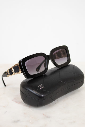 Chanel sunglasses with a black acetate frame, gold-tone chain detailing on the arms, and dark lenses, displayed on top of their signature black quilted leather case against a white marble background.