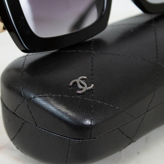 Black Chanel sunglasses with a black quilted leather case featuring a silver interlocking CC logo, displayed on a light marble surface.
