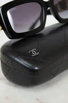 Black Chanel sunglasses with a black quilted leather case featuring a silver interlocking CC logo, displayed on a light marble surface.