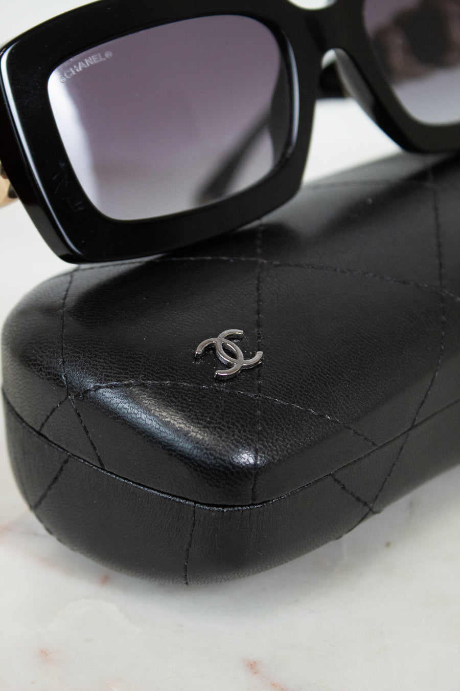 Black Chanel sunglasses with a black quilted leather case featuring a silver interlocking CC logo, displayed on a light marble surface.