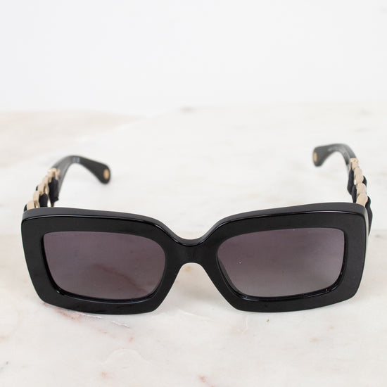 Black rectangular cat-eye sunglasses with dark lenses and gold-tone chain detailing on the arms, resting on a light marble surface.