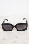 Black rectangular cat-eye sunglasses with dark lenses and gold-tone chain detailing on the arms, resting on a light marble surface.