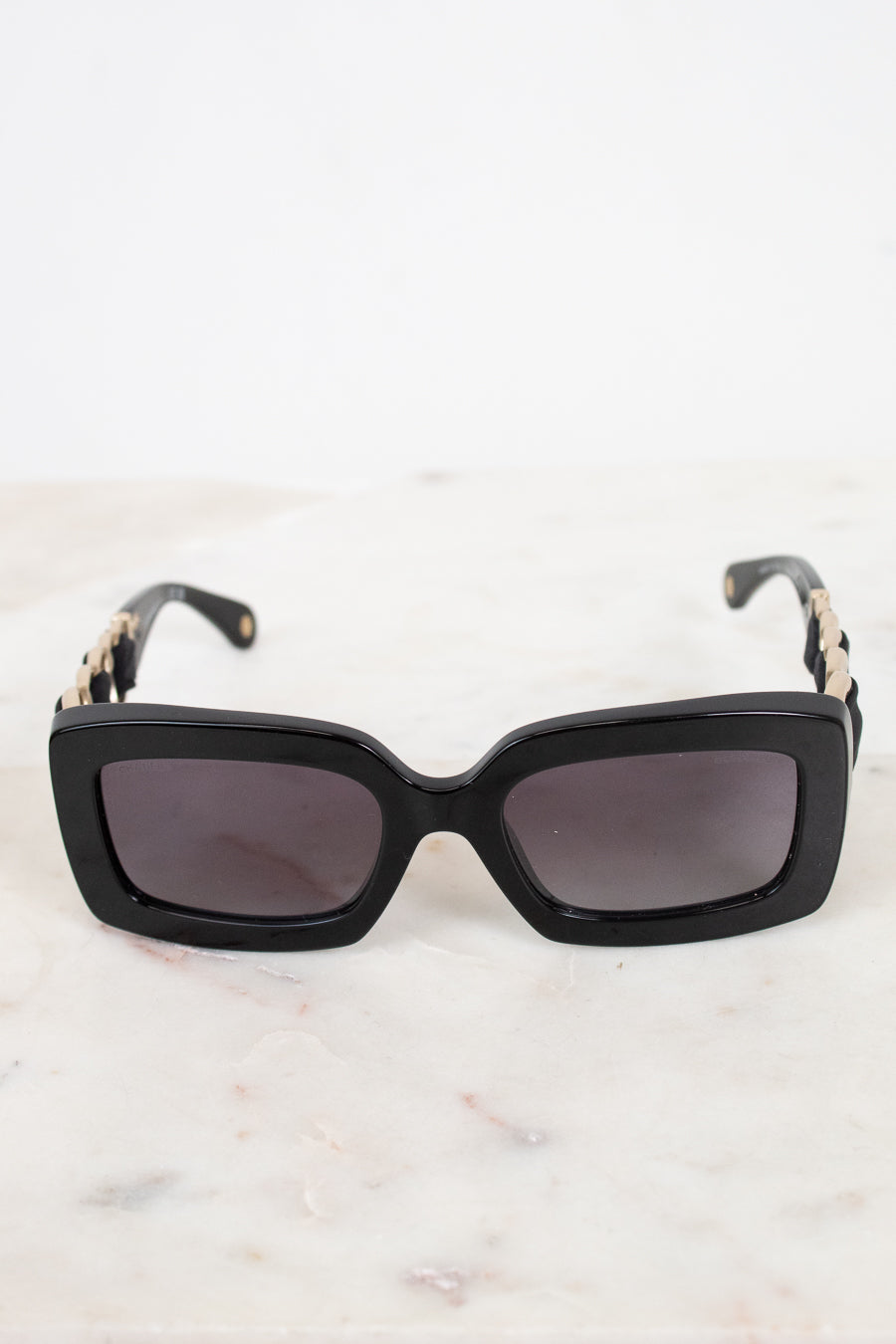 Black rectangular cat-eye sunglasses with dark lenses and gold-tone chain detailing on the arms, resting on a light marble surface.