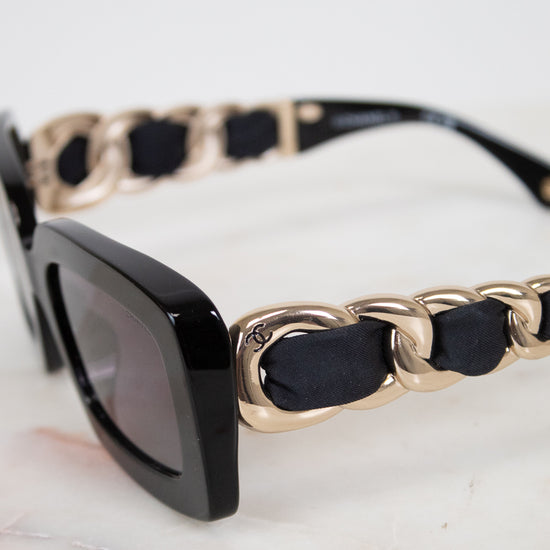Close-up of a pair of sunglasses with a glossy black acetate frame, dark gray lenses, and a unique gold-tone chain-link strap accented with black fabric woven arm details, resting on a light marble surface.