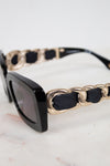 Close-up of a pair of sunglasses with a glossy black acetate frame, dark gray lenses, and a unique gold-tone chain-link strap accented with black fabric woven arm details, resting on a light marble surface.