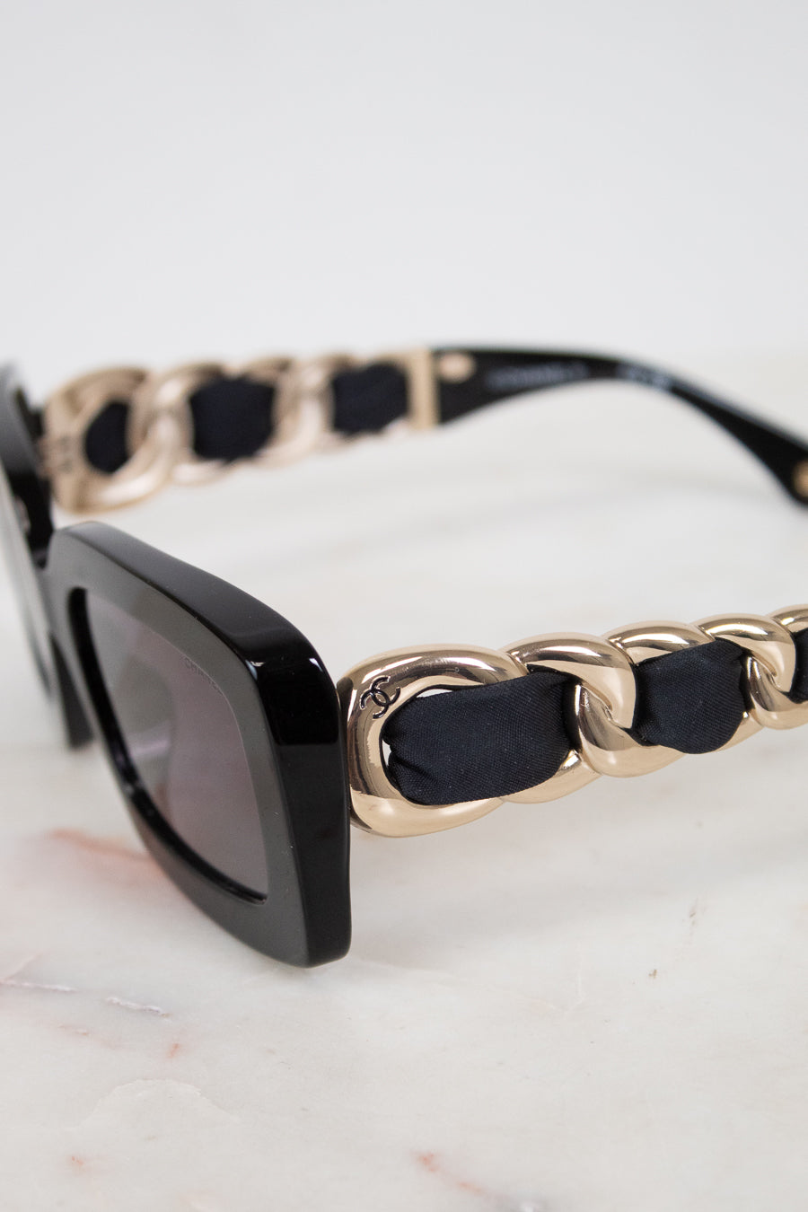Close-up of a pair of sunglasses with a glossy black acetate frame, dark gray lenses, and a unique gold-tone chain-link strap accented with black fabric woven arm details, resting on a light marble surface.