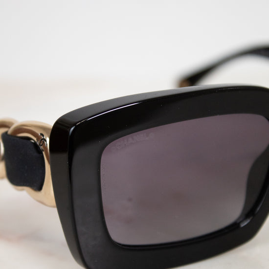 Close-up of a pair of sunglasses with a glossy black acetate frame, dark gray lenses, and gold-tone metal arms= details resting on a light marble surface.