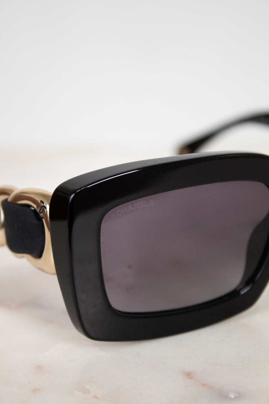 Close-up of a pair of sunglasses with a glossy black acetate frame, dark gray lenses, and gold-tone metal arms= details resting on a light marble surface.