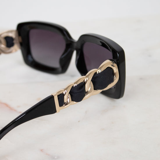 Black square sunglasses with dark lenses and chunky gold chain detailing on the arms, resting on a light marble surface.