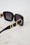 Black square sunglasses with dark lenses and chunky gold chain detailing on the arms, resting on a light marble surface.