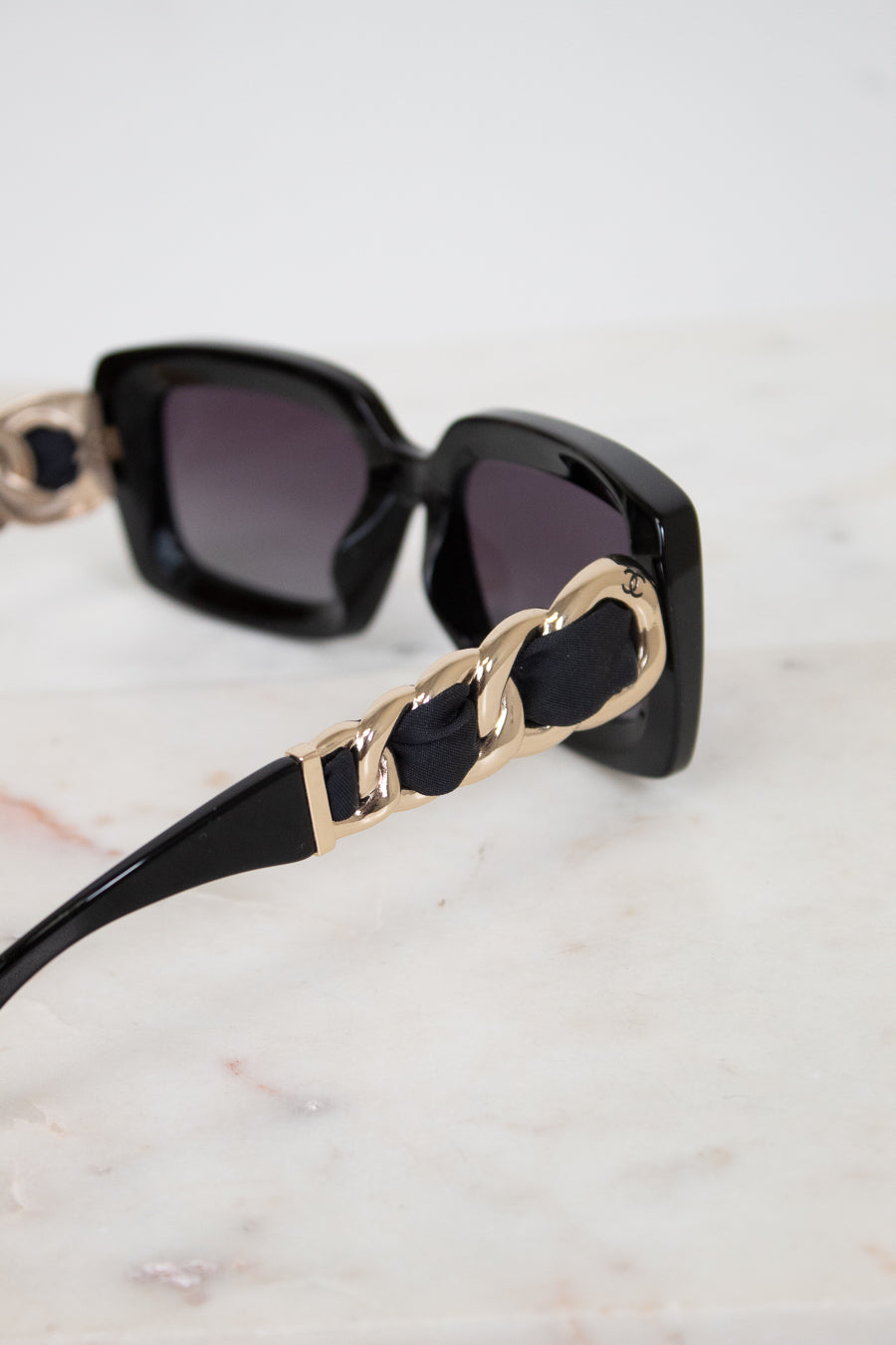 Black square sunglasses with dark lenses and chunky gold chain detailing on the arms, resting on a light marble surface.