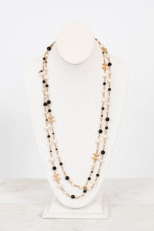 Multi-stacked necklace with black and faux pearl beads and gold-tone chain on a white stand