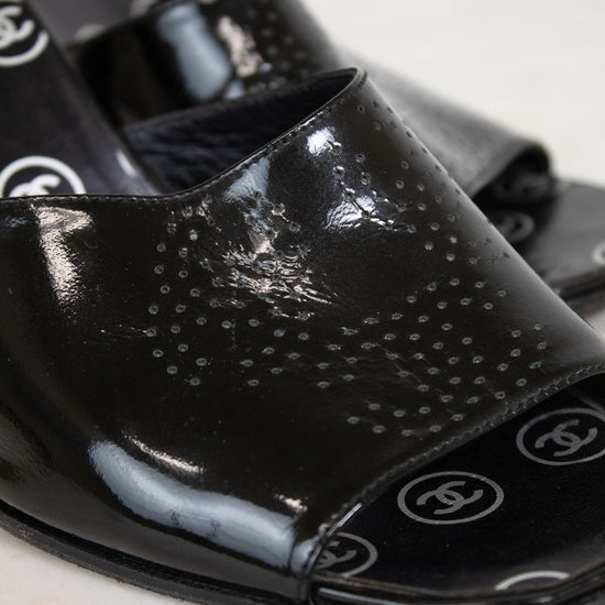 A close-up of glossy black patent leather slide sandals featuring a perforated dot pattern on the upper and CC logo details on the toe bed, set against a light marble background.