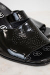 A close-up of glossy black patent leather slide sandals featuring a perforated dot pattern on the upper and CC logo details on the toe bed, set against a light marble background.