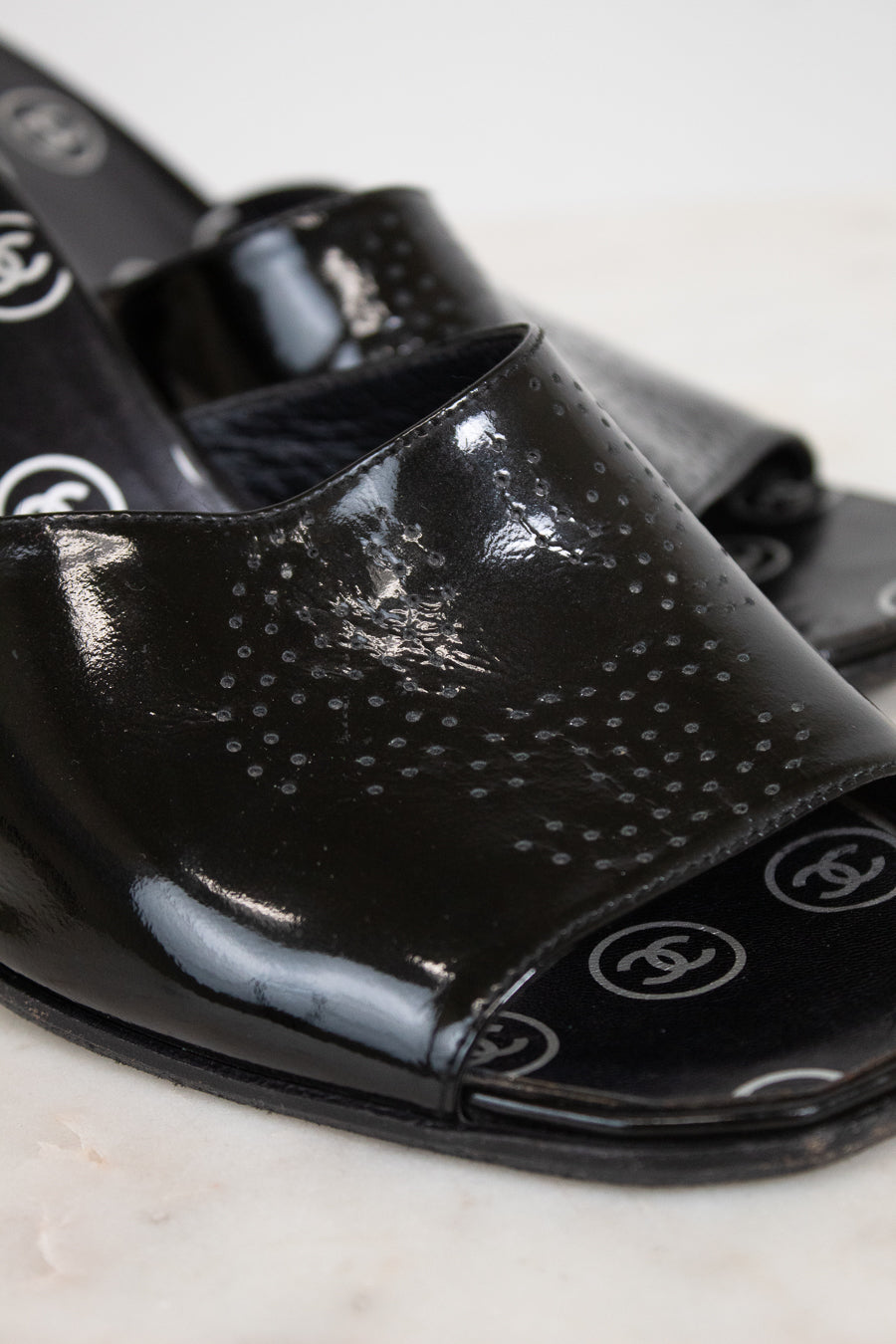 A close-up of glossy black patent leather slide sandals featuring a perforated dot pattern on the upper and CC logo details on the toe bed, set against a light marble background.
