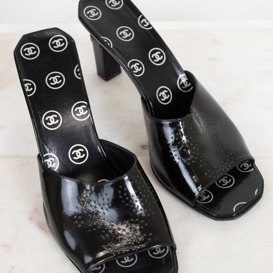 A pair of black patent leather mule sandals featuring a repeating white Chanel logo pattern on the insole and outer sole, displayed on a light marble surface.