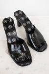A pair of black patent leather mule sandals featuring a repeating white Chanel logo pattern on the insole and outer sole, displayed on a light marble surface.