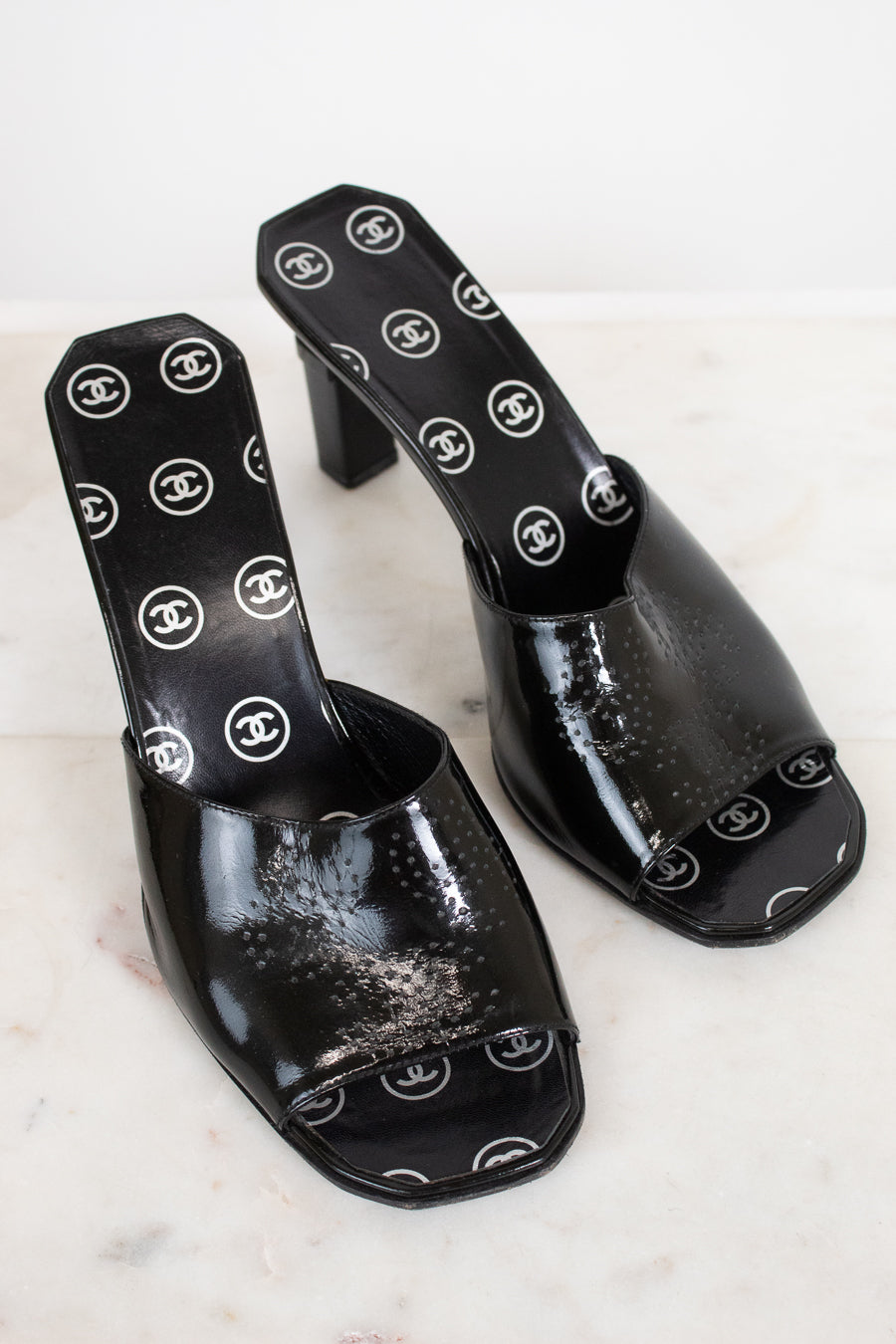 A pair of black patent leather mule sandals featuring a repeating white Chanel logo pattern on the insole and outer sole, displayed on a light marble surface.