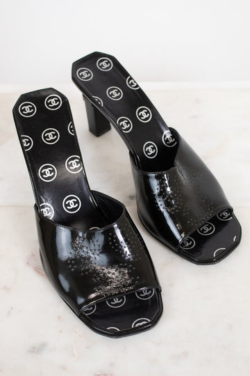 A pair of black patent leather mule sandals featuring a repeating white Chanel logo pattern on the insole and outer sole, displayed on a light marble surface.