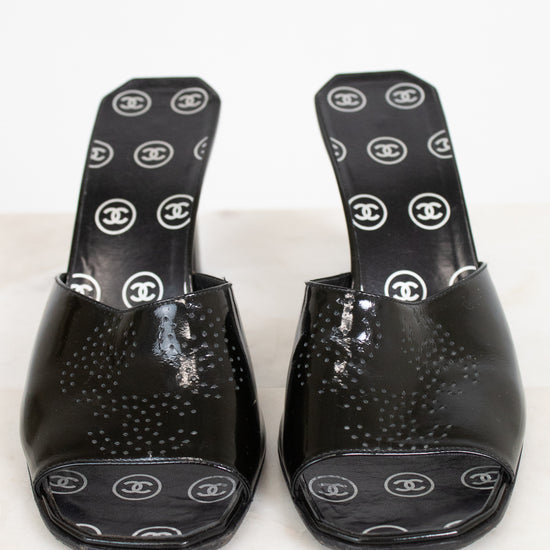 A pair of black patent leather Chanel slide mules with a textured dotted pattern on the toe area and white Chanel logo print on the insoles, displayed on a light marble surface.