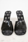 A pair of black patent leather Chanel slide mules with a textured dotted pattern on the toe area and white Chanel logo print on the insoles, displayed on a light marble surface.