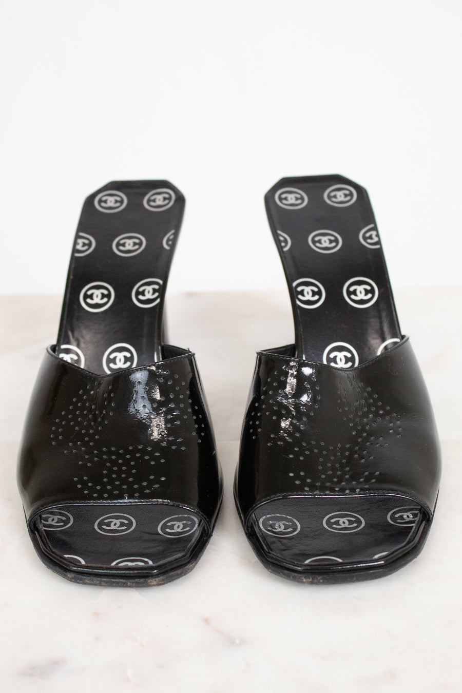 A pair of black patent leather Chanel slide mules with a textured dotted pattern on the toe area and white Chanel logo print on the insoles, displayed on a light marble surface.