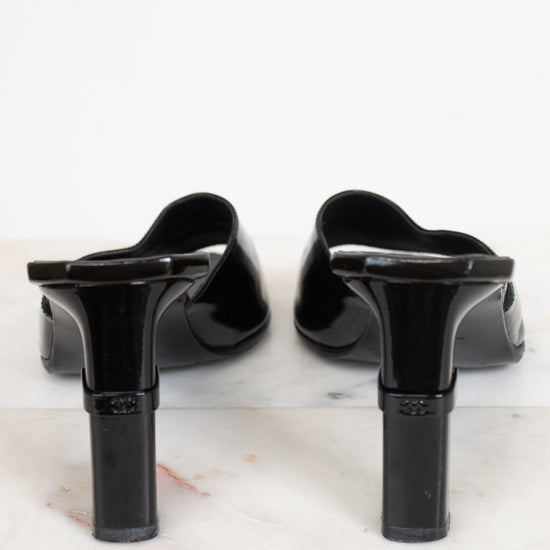 A pair of black patent leather mule heels with block heels, shown from the back on a white marble surface against a white background.