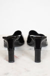 A pair of black patent leather mule heels with block heels, shown from the back on a white marble surface against a white background.