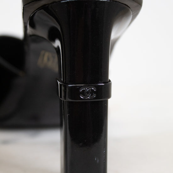 A close-up of a black patent leather high-heeled shoe heel, showing a Chanel logo band around the middle, on a white surface.